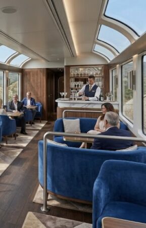 Elegant vintage‑inspired interior of a luxury train lounge car featuring polished wood panels, brass accents, and large panoramic windows looking out onto a scenic landscape.