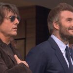 Tom Cruise and David Beckham standing side-by-side in a stadium, smiling and looking upward during the national anthem.