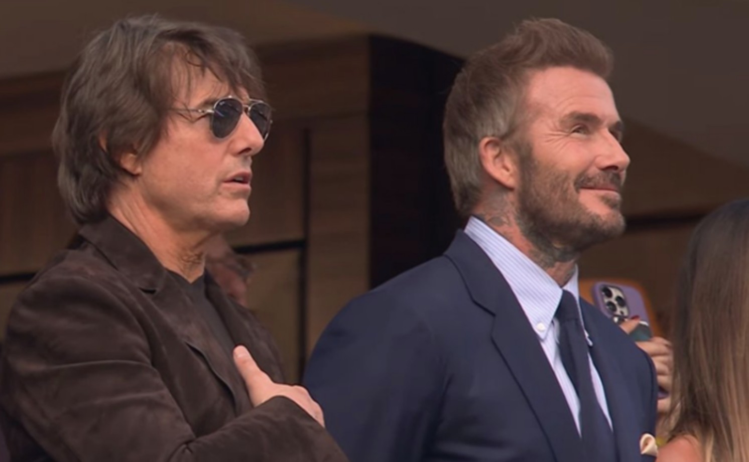 Tom Cruise and David Beckham standing side-by-side in a stadium, smiling and looking upward during the national anthem.