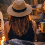 Karen Lake sits with her back to the camera wearing a straw hat and blanket, joined by two friends laughing around a glowing firepit outside The Ren cabin in Hocking Hills, Ohio. String lights and forest trees frame the cozy nighttime scene.